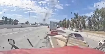 Vista de un choque en una autopista con vehículos, un camión, escombros volando, cielo con nubes y árboles. Se ve un espejo retrovisor