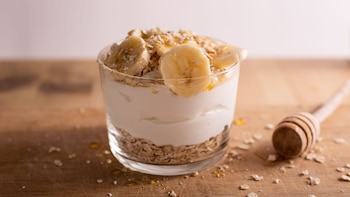 Un vaso de vidrio con helado, avena y rodajas de plátano bañadas en miel junto a un servidor de miel de madera