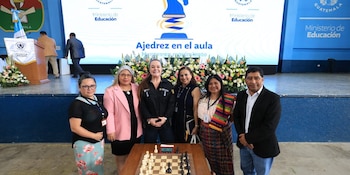 Seis personas posan detrás de un tablero de ajedrez con un reloj digital frente a una pantalla del Ministerio de Educación de Guatemala