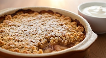 Crumble de pera y manzana con topping dorado y azúcar glas, presentado en molde de cerámica. Un tazón blanco con yogur natural se ve al costado.
