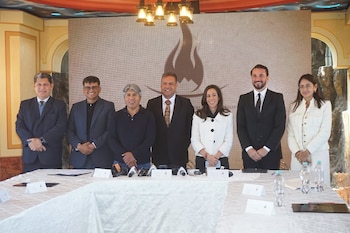 Siete personas posan detrás de una mesa con micrófonos, botellas de agua y documentos, frente a una pantalla grande con un logotipo de llama