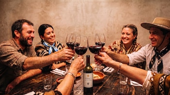 Varias personas alzan copas de vino tinto sobre una mesa de madera donde hay una botella de vino y platos.