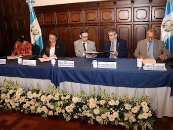 Cinco funcionarios sentados ante una mesa azul firman documentos frente a dos banderas de Guatemala y arreglos de flores blancas