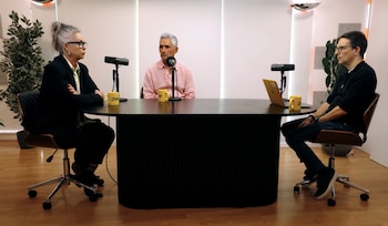 Tres personas sentadas en sillas de oficina alrededor de una mesa negra. Hay dos micrófonos, un ordenador portátil amarillo y dos tazas amarillas con el logo Zoe