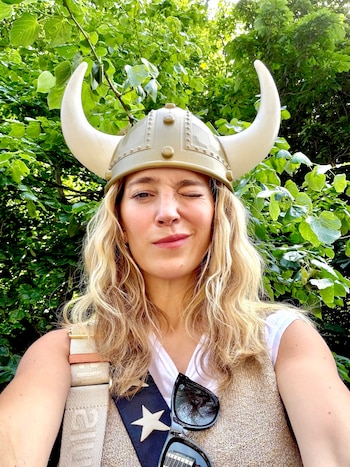 Retrato cercano de mujer rubia con cabello largo, casco vikingo con cuernos, camiseta blanca, chaleco beige y gafas de sol, guiñando un ojo. Fondo verde de hojas