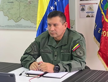 Hombre con uniforme militar verde y parche de la bandera de Venezuela sentado a una mesa con libreta, bolígrafo y tableta electrónica