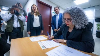 Una mujer con lentes y cabello rizado firma un papel sobre una mesa de madera mientras otras personas la observan