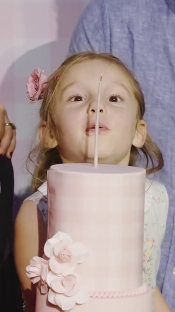 Niña con flor rosa en el pelo frente a una torta rosa con flores y una vela encendida. Brazo de adulto visible al costado