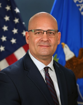 Retrato oficial de un hombre con anteojos, traje oscuro y corbata, ubicado frente a la bandera de Estados Unidos