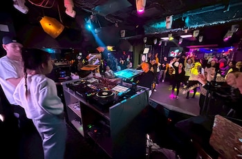 Una cabina de DJ con mesa de mezclas y computadora frente a varias personas que bailan bajo luces de colores en una discoteca