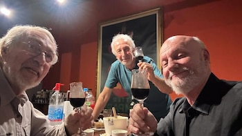 Tres hombres, Luis Brandoni, Eduardo Blanco y Juan José Campanella, sonríen y brindan con copas de vino tinto frente a una pared roja con un cuadro