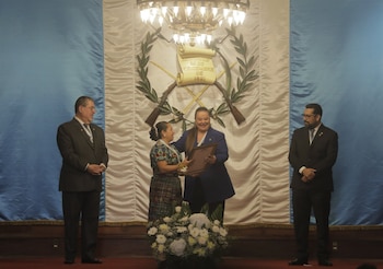 Cuatro personas en un escenario con la bandera de Guatemala y su escudo. Dos mujeres en el centro, una entrega un marco a la otra. Arreglo floral y candelabro