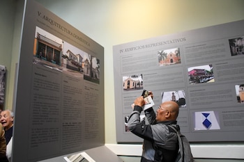 Un hombre calvo con gafas fotografía paneles de exhibición con imágenes y textos sobre arquitectura. Otro hombre asoma por la izquierda. Luz cenital