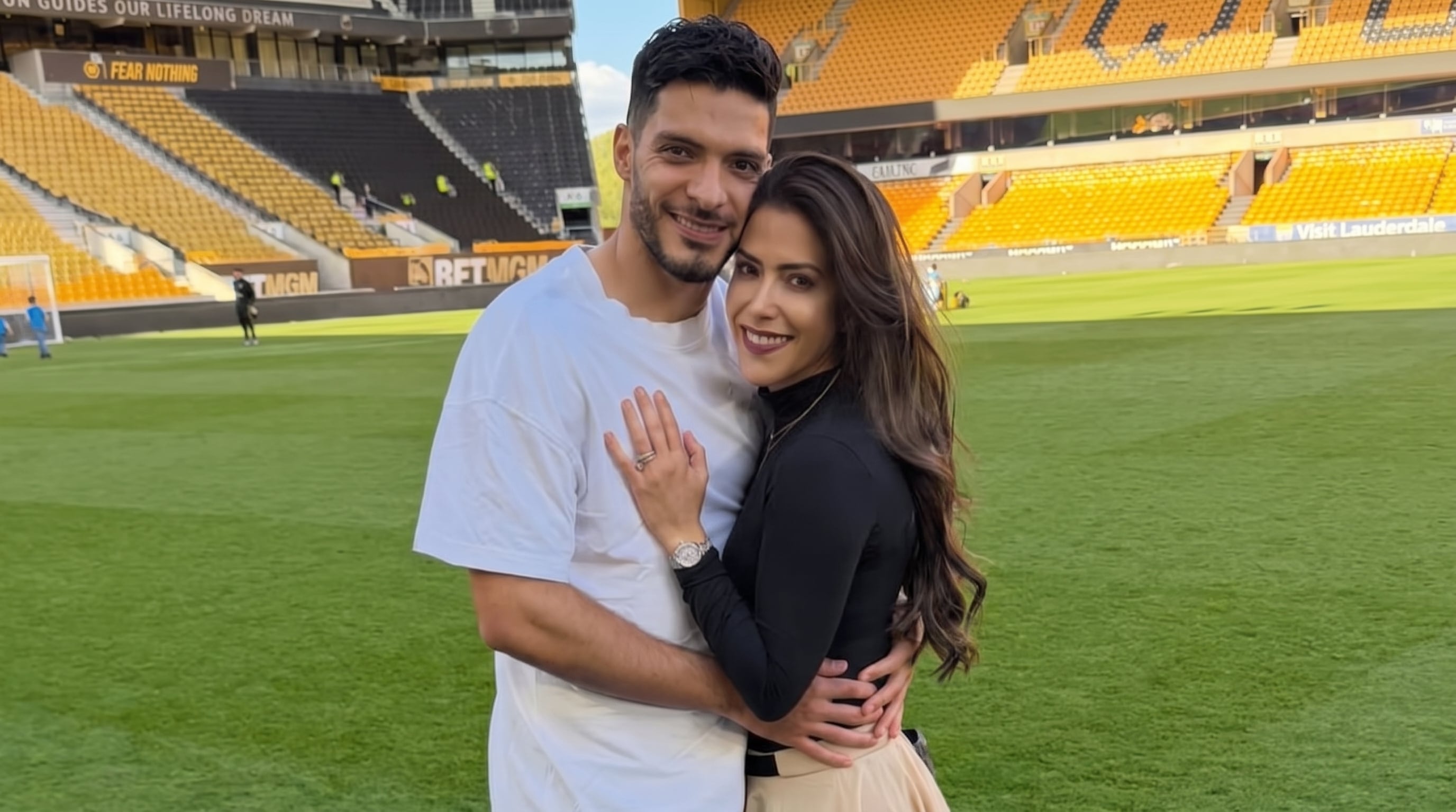 Daniela Basso y Raúl Jiménez posan abrazados en el campo de un estadio de fútbol con las gradas al fondo. (Imagen Ilustrativa Infobae)