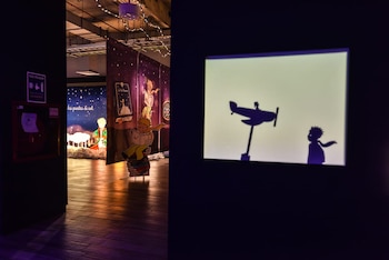 Sala de exposición con recortes del Principito, paneles ilustrados de la obra, un extintor de incendios y una proyección de siluetas de un avión y el niño