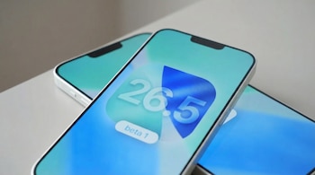 Primer plano de un smartphone con pantalla iluminada, mostrando "26.5 beta 1" sobre un fondo degradado azul y turquesa; se aprecian otros dos teléfonos detrás.