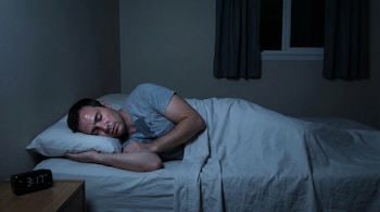 Hombre durmiendo de lado en cama con sábanas claras. Su rostro y cuello están sudorosos. Viste camiseta gris. Reloj digital marca 3:17 AM en mesita. Habitación oscura.