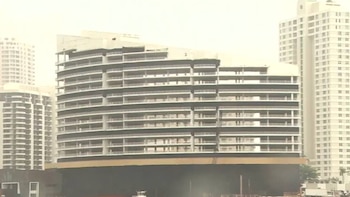 Vista frontal de um grande módulo curvo de um edifício de vários andares com varanda em uma plataforma escura na água, com arranha-céus ao fundo.