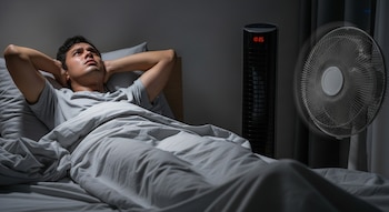 Un hombre con una camiseta gris yace en la cama bajo una sábana ligera, con sudor en la frente, mientras un ventilador de pie oscila a su lado.