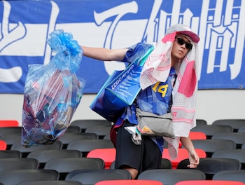 Hombre con gorra, gafas y bufanda, recogiendo basura en una bolsa plástica transparente entre asientos de estadio. Al fondo, asientos y una bandera azul