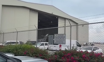 Un hangar con puerta abierta muestra un avión pequeño detrás de una valla metálica con alambre de púas y un cartel de prohibido el paso. Varios vehículos y arbustos
