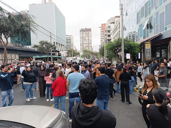 Una calle urbana está llena de personas y vehículos detenidos. Se observan edificios de gran altura y árboles a lo largo de la vía