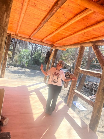 Una mujer de cabello corto y camiseta blanca sonríe bajo un porche de madera con vigas rústicas y piso de madera. Hay árboles al fondo y mucha luz solar