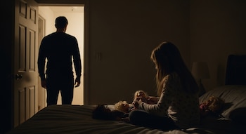 Un hombre de espaldas en el umbral de una puerta abierta y una niña de espaldas sentada en la cama juega con tres muñecas, todo en una habitación oscura.