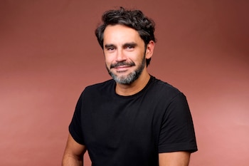 Retrato de un hombre de mediana edad, Gonzalo Heredia, con cabello oscuro rizado y barba, vestido con una camiseta negra simple sobre un fondo marrón rojizo liso