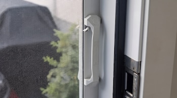 Ventana con mosquitero negro, marco blanco, asa blanca con pestillo metálico, bisagra y follaje verde borroso al fondo.