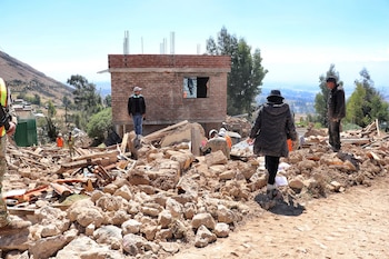 Terreno cubierto de rocas y escombros, un edificio de ladrillo parcial, varias personas caminando, montaña al fondo bajo un cielo azul claro