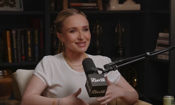 Hayden Panettiere sonríe y gesticula con las manos en un estudio. Viste una blusa blanca, collares dorados, con un micrófono de podcast al frente y estanterías detrás