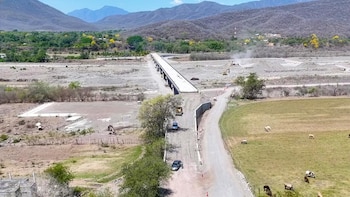 Longa ponte construída sobre um rio seco com leito de pedra, rodeada de vegetação e montanhas. Ladrões, carros e animais são vistos