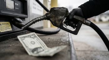 Una mano enguantada sostiene una boquilla de surtidor de combustible junto a una bomba de gasolina. Un billete de un dólar estadounidense desenfocado reposa en la base.