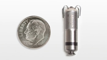 Un marcapasos Medtronic Micra cilíndrico, plateado y blanco con ganchos, al lado de una moneda de diez centavos de dólar estadounidense sobre un fondo blanco