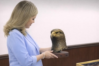 Una mujer rubia con un blazer azul claro y un brazalete de perlas sostiene una escultura de cabeza de águila en bronce sobre una base de madera oscura.