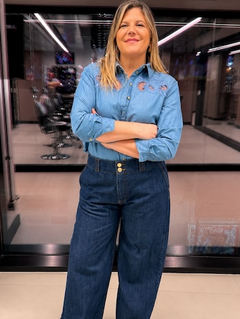 Dominique Metzger, de mediana edad, de pie, sonriendo, brazos cruzados. Viste camisa y pantalón de mezclilla azul. Fondo de vidrio con reflejos