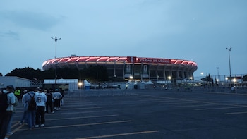 Vista exterior del Estadio Ciudad de México al anochecer, iluminado con luces rojas y mostrando banners del Mundial 2026. Un grupo de personas se ve en un estacionamiento