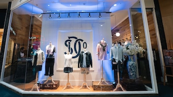 Vitrina comercial con seis maniquíes vestidos con ropa casual, maletas antiguas en el piso y un jarrón con flores blancas