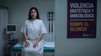 Una mujer joven con cabello oscuro y bata de hospital está sentada en una camilla de examen médico. A su izquierda hay un monitor y a su derecha un cartel púrpura borroso.