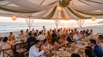 Banquete de boda en playa salvadoreña. Invitados sentados en mesas bajo una carpa blanca decorada con telas y lámparas, con el Océano Pacífico y un atardecer dorado.