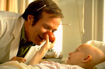 Hombre con bata blanca, nariz de payaso roja, hisopos en el cabello, sonríe a un niño calvo sonriente acostado en una cama de hospital