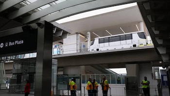 Un tren blanco elevado se desplaza sobre una estructura de estación moderna. Abajo, se ven trabajadores con chalecos amarillos y un cartel de 'Bus Plaza'