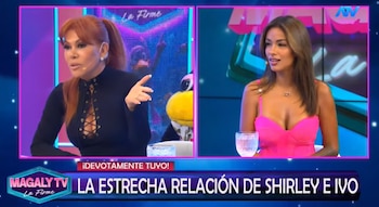 Dos mujeres sentadas en un estudio de televisión frente a pantallas, una de cabello rojizo con top negro, otra de cabello castaño con top fucsia, un títere amarillo a un lado