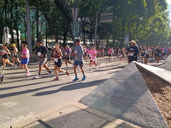 Muitos corredores caminham pelas ruas arborizadas da Cidade do México, vestindo babadores, camisas esportivas e prédios ao fundo.