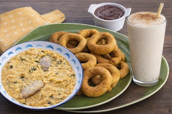 Plato de mazamorra, rosquillas, vaso de bebida con canela y pajita, cuenco con salsa oscura, servilleta amarilla y mesa de madera.