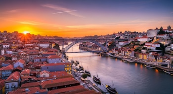 Vista aérea de la ciudad de Oporto al atardecer, mostrando edificios, el río Duero con barcos, y el puente Dom Luís I. El sol se oculta en el horizonte con resplandor.