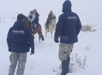 Dos agentes de policía con uniformes oscuros, vistos de espaldas, caminan sobre la nieve. Tres personas montan dos caballos al fondo