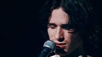 Primer plano de Jeff Buckley, hombre de cabello oscuro y desordenado, cantando en un micrófono. Su rostro está iluminado con los ojos ligeramente cerrados, sobre un fondo oscuro