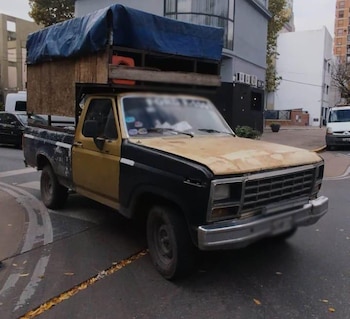 Camioneta pickup Chevrolet bicolor (amarillo ocre y negro) con carrocería de madera y lona azul sobre la caja. Estacionada en una calle urbana con edificios